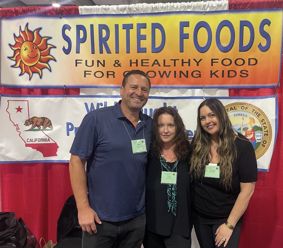 Three people smiling at a Spirited Foods booth promoting healthy snacks for kids.